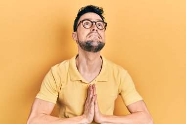 Gündelik kıyafetler ve gözlük takan genç İspanyol adam elleri ile yalvarıyor ve dua ediyor. Yüzünde umut ifadesi var. Çok duygusal ve endişeli. Yalvarıyor. 
