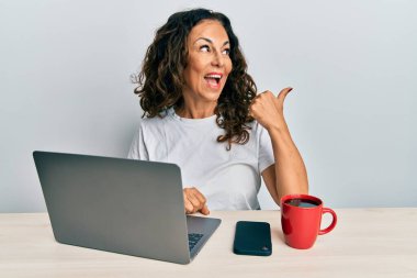 Ofiste çalışan orta yaşlı güzel bir kadın bilgisayarlı dizüstü bilgisayar kullanıyor. Gülümsüyor, gülümsüyor ve başparmağıyla yan tarafa bakıyor.. 