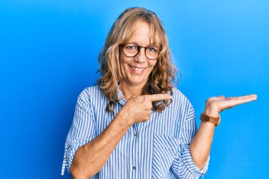 Orta yaşlı, sıradan kıyafetler ve gözlük takan sarışın kadın kameraya hayret ediyor ve gülümsüyor.. 