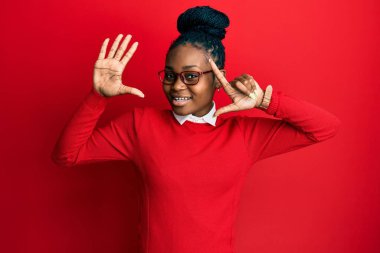 Genç Afro-Amerikalı kadın günlük kıyafetler ve gözlük takıyor. Yedi numaralı parmaklarıyla işaret ediyor. Kendinden emin ve mutlu gülümsüyor.. 