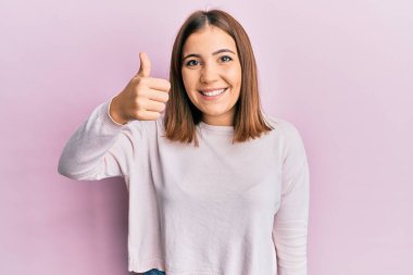 Genç ve güzel bir kadın günlük kıyafetler giyip gülümsüyor mutlu ve pozitif, başparmağıyla mükemmel ve onay işareti yapıyor. 