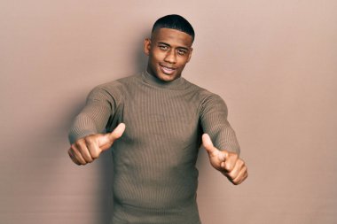 Balıkçı yaka kazak giyen genç siyahi adam olumlu bir jest yapmayı onaylıyor. Elleriyle gülümsüyor ve başarı için seviniyor. kazanan jest. 