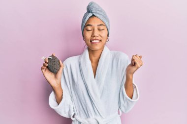 Young chinese woman wearing shower towel cap and bathrobe holding pumice stone screaming proud, celebrating victory and success very excited with raised arm 