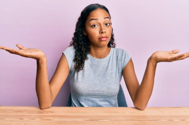 Gündelik giysiler giymiş genç Afro-Amerikalı kız masanın üzerinde elleri ve kolları havada, kafası karışmış bir şekilde oturuyor. şüphe kavramı. 