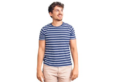 Kıvırcık saçlı, sıradan kıyafetler giyen, yüzünde gülümseme, doğal bir ifade olan genç yakışıklı bir adam. Kendinden emin gülen. 