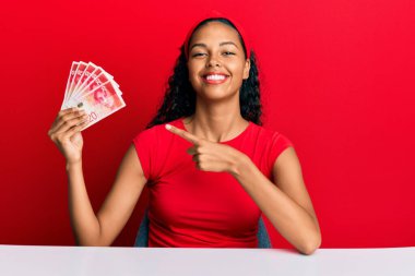 Genç Afrikalı Amerikalı kız İsrail şekellerini tutuyor masada oturuyor mutlu bir şekilde el ve parmaklarıyla işaret ediyor 