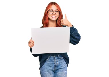 Boş afiş taşıyan genç kızıl saçlı kadın mutlu ve olumlu gülümsüyor, başparmağıyla mükemmel ve onay işareti yapıyor. 