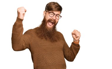 Genç İrlandalı kızıl adam günlük kıyafetler ve gözlük takıyor mutlu ve neşeli dans ediyor, gülümsüyor rahat ve kendinden emin bir şekilde müzik dinliyor. 