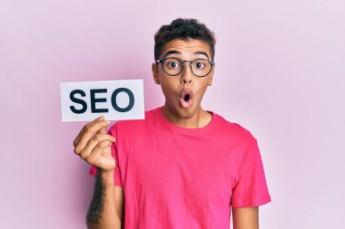 Young handsome african american man holding seo message paper scared and amazed with open mouth for surprise, disbelief face 