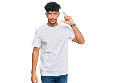 Gündelik beyaz tişört giyen genç Arap adam gülümsüyor ve kendinden emin el hareketleriyle küçük beden işareti yapıyor parmaklarıyla bakıyor ve kameraya bakıyor. ölçüm kavramı. 