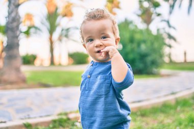 Üzgün küçük çocuk, güneşli bir günde parkta diş etlerine dokunduğu için parmaklarını dudaklarına koyuyor. Güzel sarı saçlı erkek bebek yeni bebek dişleri için acı çekiyor.