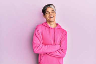 Sıradan pembe kazak giyen genç İspanyol adam kollarını kavuşturup kameraya bakarken gülümsüyor. pozitif kişi. 