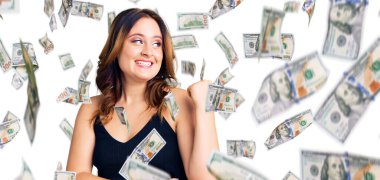 Genç, güzel, beyaz, günlük kıyafetler giyen mutlu yüzüyle gülümseyen ve baş parmağıyla yüzünü gösteren bir kadın..