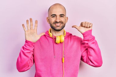 Young bald man wearing gym clothes and using headphones showing and pointing up with fingers number six while smiling confident and happy. 