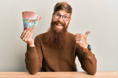 Genç İrlandalı kızıl saçlı adam elinde 100 yeni Zelanda Doları banknotuyla masanın üzerinde oturmuş başparmağıyla yan tarafı işaret ederek mutlu bir şekilde gülümsüyor. 