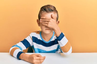 Kafkasyalı sevimli bir çocuk, gündelik giysiler giyip masaya oturmuş, gözlerini eliyle kapatmış, ciddi ve üzgün görünüyor. Görme, saklama ve reddetme konsepti 