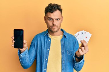 Poker kartları ve akıllı telefon tutan genç İspanyol bir adam boş ekran şüphecisi ve gergin görünüyor, sorun yüzünden suratını asıyor. negatif kişi. 