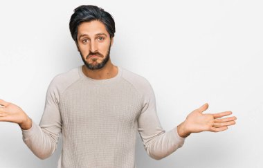 Sıradan kıyafetler giyen, elleri ve kolları havada, kafası karışmış genç İspanyol bir adam. şüphe kavramı. 