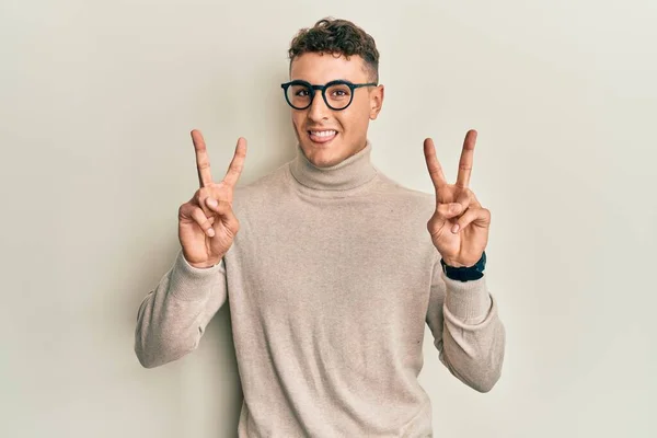 스페인 청년은 터틀넥 스웨터를 웃으면서 손가락으로 승리의 표시를 — 스톡 사진