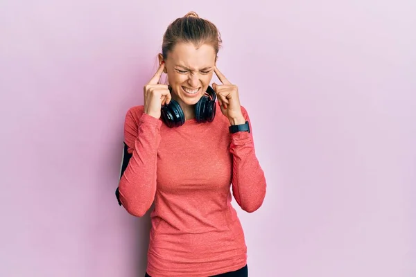 Beautiful caucasian woman wearing sportswear and arm band covering ears with fingers with annoyed expression for the noise of loud music. deaf concept.