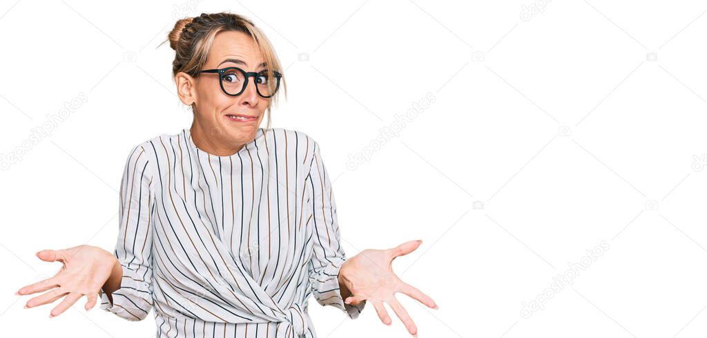 Beautiful blonde woman wearing business shirt and glasses clueless and confused expression with arms and hands raised. doubt concept. 