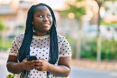 Genç Afrikalı Amerikalı kadın şehirde akıllı telefon kullanarak mutlu bir şekilde gülümsüyor..