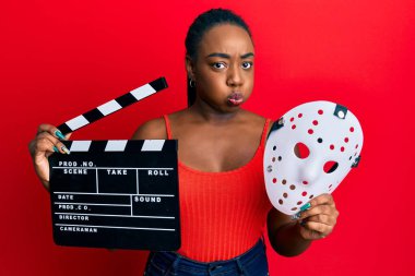 Young african american woman holding video film clapboard and fear mask puffing cheeks with funny face. mouth inflated with air, catching air. 