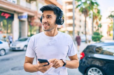 Genç Latin adam şehirde akıllı telefon ve kulaklık kullanarak mutlu bir şekilde gülümsüyor..