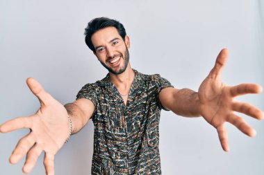 Zarif bir yaz gömleği giyen genç İspanyol adam kameraya bakıp sarılmak için kollarını açmış gülümsüyor. Mutluluğu kucaklayan neşeli bir ifade.. 