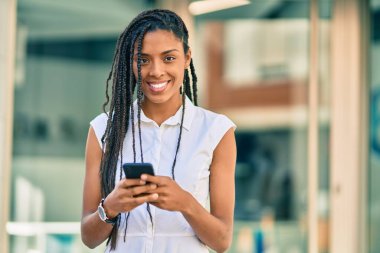 Genç Afrikalı Amerikalı kadın şehirde akıllı telefon kullanarak mutlu bir şekilde gülümsüyor..