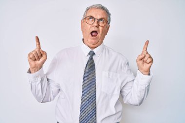 İş kıyafetleri giyen yaşlı gri saçlı adam şaşkın şaşkın yukarı bakıyor ve parmaklarıyla işaret ediyor ve kollarını kaldırıyor.. 