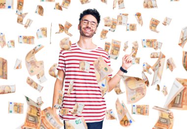 Yüzünde kocaman bir gülümseme olan, gündelik kıyafetler giyen genç İspanyol adam, kameraya el ile işaret ediyor..