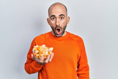 Young bald man holding potato chip scared and amazed with open mouth for surprise, disbelief face 