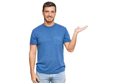 Yakışıklı, günlük kıyafetler giyen, neşeli bir şekilde gülümseyen, avuç içiyle kameraya bakan beyaz bir adam.. 