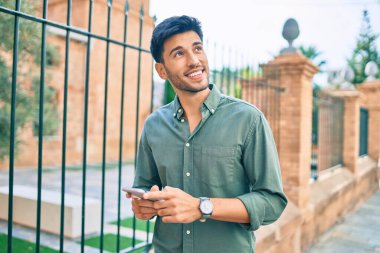 Genç Latin adam mutlu bir şekilde gülümsüyor ve şehirde akıllı telefon kullanıyor..