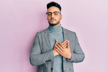 İş kıyafetleri giymiş genç bir Arap, elleri göğsünde gülümsüyor, gözleri kapalı, yüzünde minnet ifadesi var. sağlık kavramı. 