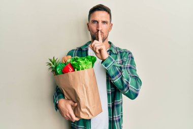 Elinde poşetlerle sakallı yakışıklı bir adam dudakları parmaklarıyla sessiz olmak istiyor. sessizlik ve gizli konsept. 