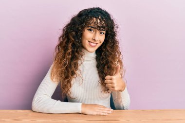 Latin asıllı genç bir kız, gündelik giysiler giyip masada oturmuş, mutlu mesut el hareketi yapıyor. Kameraya bakıp başarıyı gösteren ifade. 