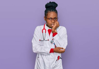 Doktor üniforması ve steteskop giyen genç Afro-Amerikan kadın elleri ağızda tırnak yerken gergin ve stresli görünüyor. anksiyete. 