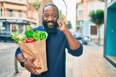 Elinde süpermarketten alınmış kağıt torba sakallı Afrikalı Amerikalı adam telefonda konuşuyor.