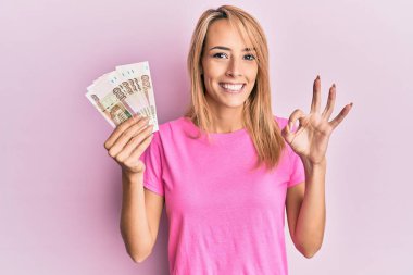 Güzel sarışın kadın elinde 100 Rus rublesi tutuyor. Tamam işareti yapıyor. Parmaklarıyla imzalıyor. 