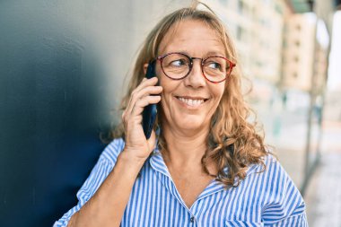 Orta yaşlı, beyaz, gülümseyen bir kadın. Şehirdeki akıllı telefondan mutlu bir şekilde konuşuyor..