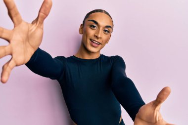 Hispanic man wearing make up and long hair wearing casual clothes looking at the camera smiling with open arms for hug. cheerful expression embracing happiness. 