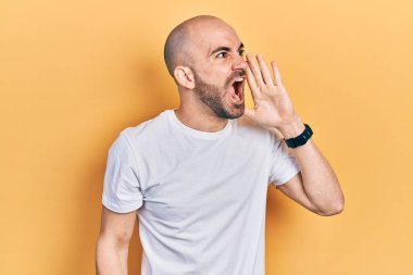 Young bald man wearing casual white t shirt shouting and screaming loud to side with hand on mouth. communication concept. 