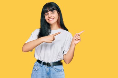 Gündelik kıyafetler giyen, gülümseyen ve kameraya bakan genç İspanyol kız iki eliyle ve parmaklarıyla kenarı işaret ediyor.. 