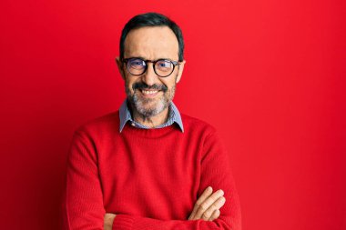 Orta yaşlı İspanyol bir adam, günlük kıyafetler ve gözlüklü mutlu bir yüz takmış, kollarını kavuşturup kameraya bakıyor. pozitif kişi. 