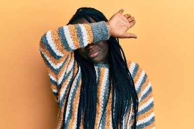 Örgülü genç siyahi bir kadın, sıradan bir kazak giyiyor, kollarıyla gözlerini kapatıyor, ciddi ve üzgün görünüyor. Görme, saklama ve reddetme konsepti 