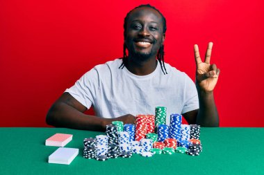 Poker fişleri ve kartlarıyla masada oturan yakışıklı, genç siyahi adam kameraya gülümseyip zafer işareti yapıyor. İki numara.. 