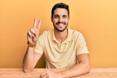 Gündelik kıyafetler giyen genç İspanyol adam masaya oturmuş gülümsüyor kameraya göz kırparak zafer işareti yapıyor. İki numara.. 