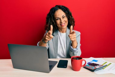Ofiste çalışan orta yaşlı güzel bir kadın bir fincan kahve içip mutlu ve komik yüzüyle kamerayı işaret ediyor. iyi enerji ve titreşimler. 